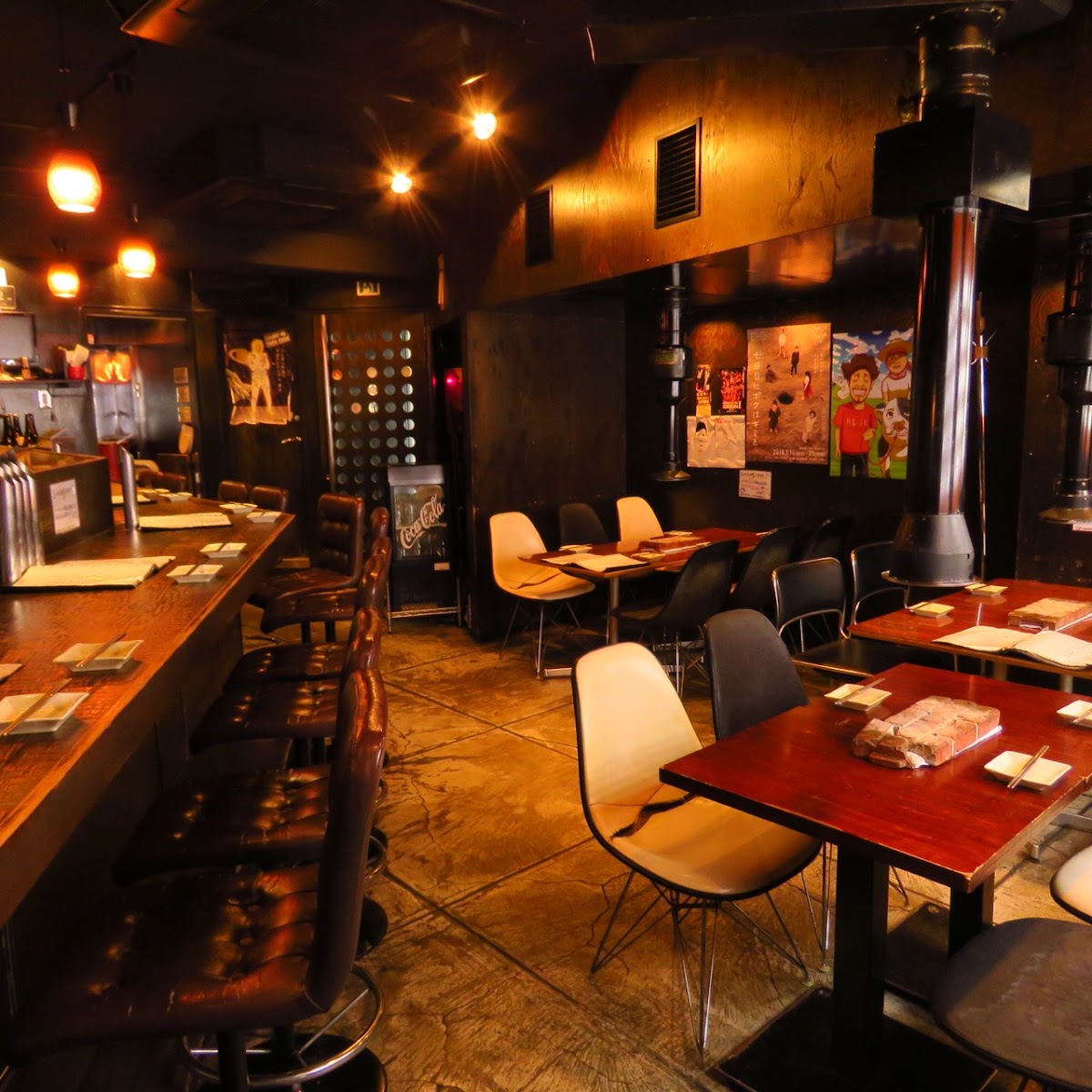 照明を落とした店内。無垢板のカウンターに背の高い革張りの椅子が並び、奥にはテーブル席。床は目地の入ったコンクリート。天井から銀色の排煙ダクトがおりている。