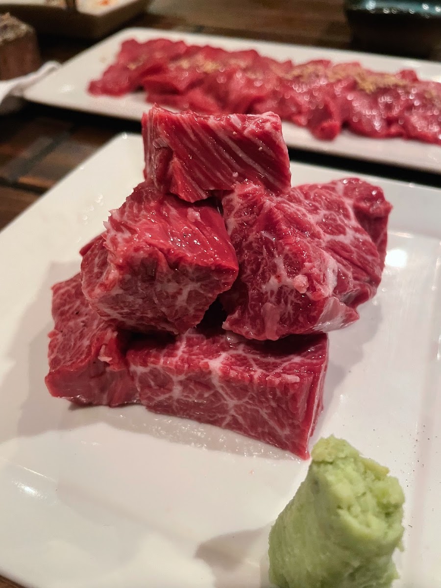 白い長皿に、さいの目に切られた牛肉のかたまりが積まれている。脂が細かく網目のように入っている。奥の皿には薄切りの肉が並び、手前にはわさびがひとつまみ添えてある。