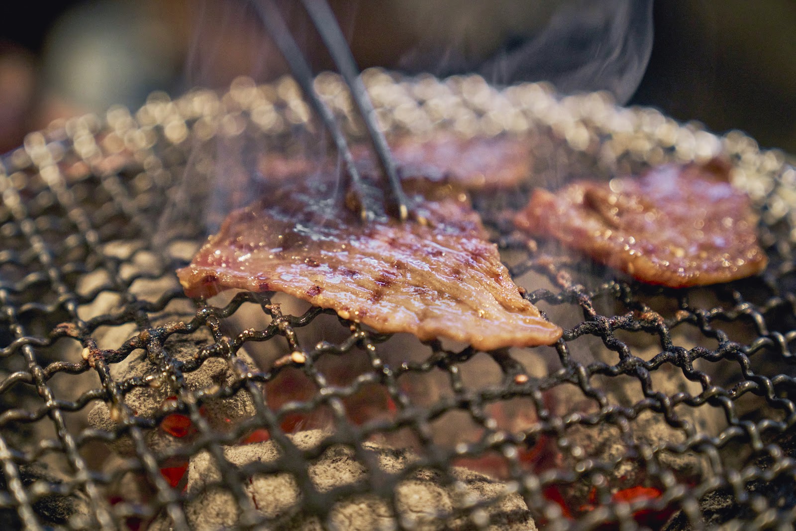 七輪の網の上で、牛肉の薄切りが一枚そりかえっている。網の下では炭が赤くおこり、白い煙が立ちのぼっている。菜箸が肉に添えられている。