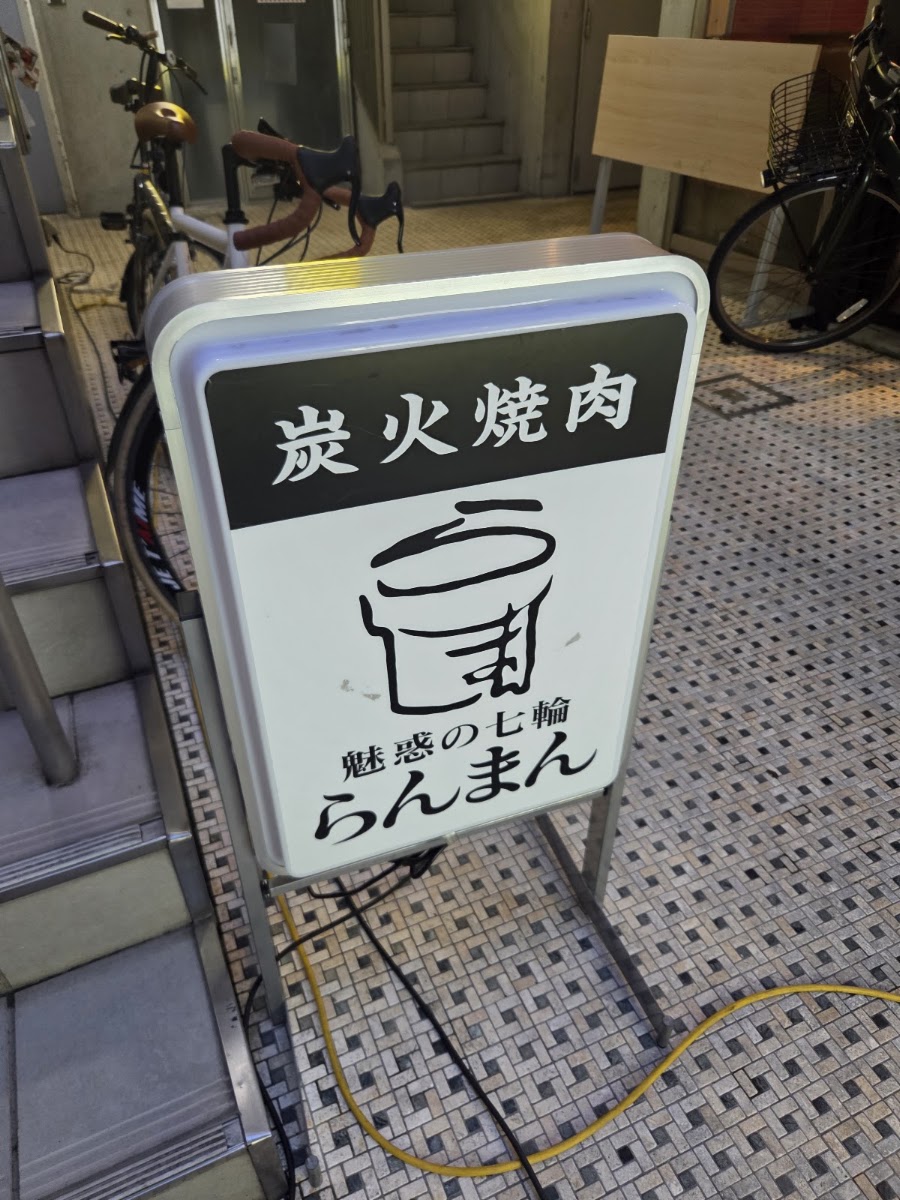 歩道に出された電飾のA型看板。上の黒帯に白抜きで「炭火焼肉」、下の白地に七輪の絵と「魅惑の七輪 らんまん」。うしろに建物の階段と自転車が写っている。